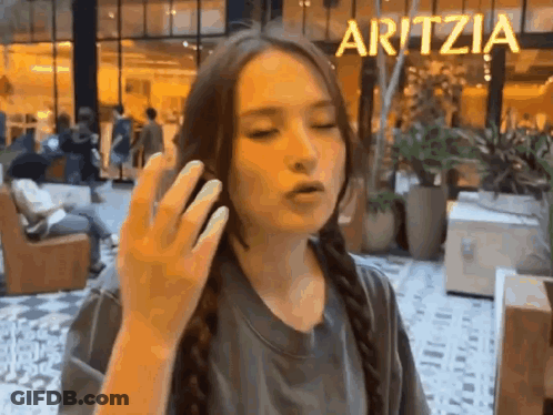 A Woman Is Talking On A Cell Phone In Front Of A Sign That Says Aritzea . GIF