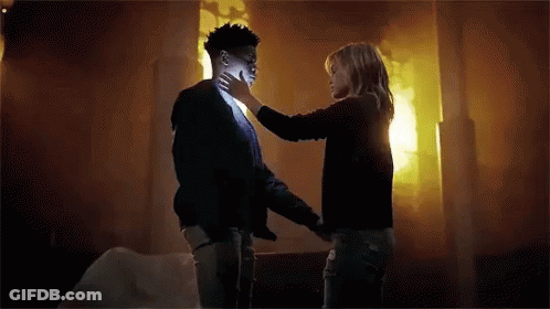 A Woman Is Touching A Man 'S Face In A Room With A Fire In The Background . GIF