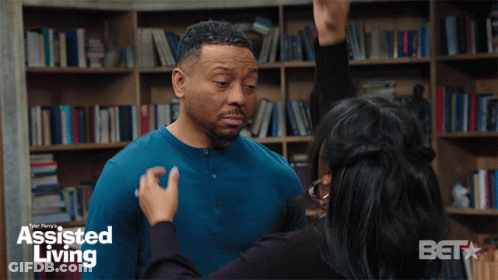 A Woman Is Touching A Man 'S Face In Front Of A Bookshelf With Assisted Living Written On It GIF