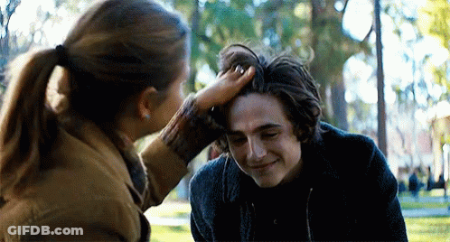 A Woman Is Touching A Young Man 'S Head In A Park . GIF