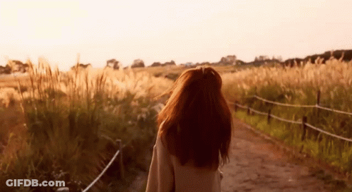 A Woman Is Walking Down A Dirt Road In A Field . GIF