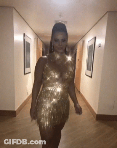 A Woman Is Walking Down A Hallway Wearing A Gold Dress GIF