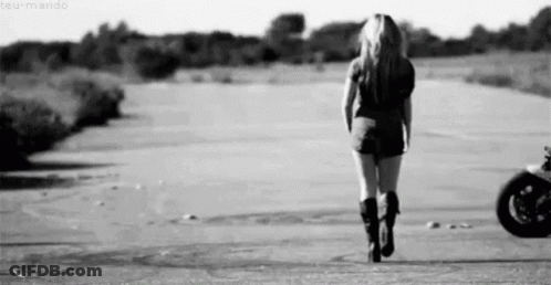 A Woman Is Walking Down A Road In Front Of A Motorcycle . GIF