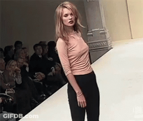 A Woman Is Walking Down A Runway At A Fashion Show Wearing A Pink Shirt And Black Pants . GIF