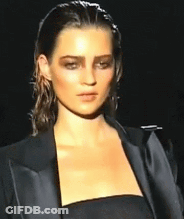 A Woman Is Walking Down A Runway Wearing A Black Jacket And A Black Dress . GIF