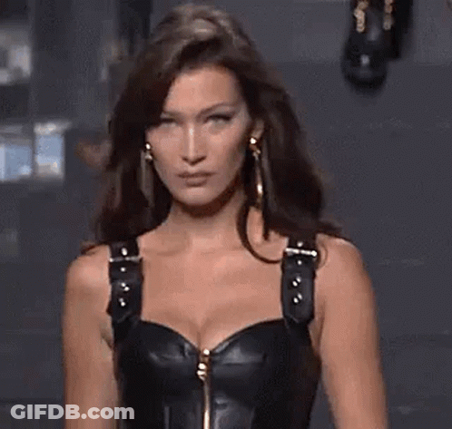 A Woman Is Walking Down A Runway Wearing A Black Leather Corset And Earrings . GIF