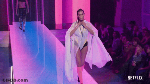 A Woman Is Walking Down A Runway Wearing A White Cape And Pink Socks . GIF