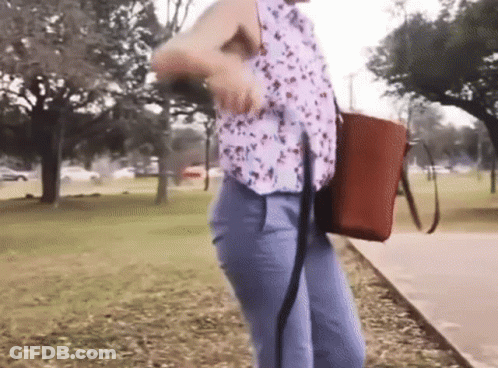 A Woman Is Walking Down A Sidewalk In A Park With A Brown Bag GIF