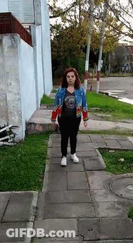 A Woman Is Walking Down A Sidewalk Wearing A Blue Jacket With Flames On The Sleeves GIF
