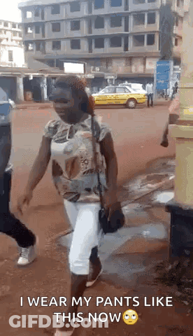 A Woman Is Walking Down A Street Wearing White Pants And A Shirt . GIF