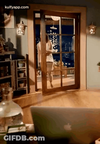 A Woman Is Walking Through A Sliding Glass Door In A Living Room Next To An Apple Laptop . GIF