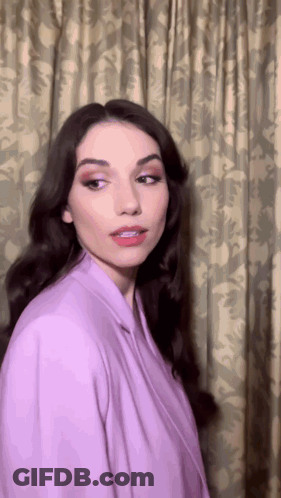 A Woman Is Wearing A Purple Jacket And Standing In Front Of A Curtain GIF