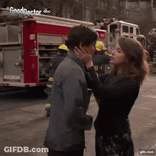 A Woman Kisses A Man On The Cheek In Front Of A Fire Truck That Says The Good Doctor On It GIF