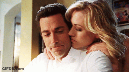 A Woman Kisses A Man On The Cheek While He Looks Down GIF