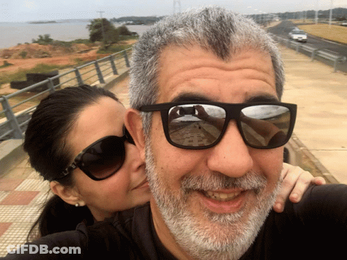A Woman Kisses A Man 'S Cheek While Wearing Sunglasses GIF