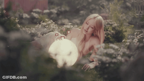 A Woman Laying In A Field Of Flowers Holding A Light GIF