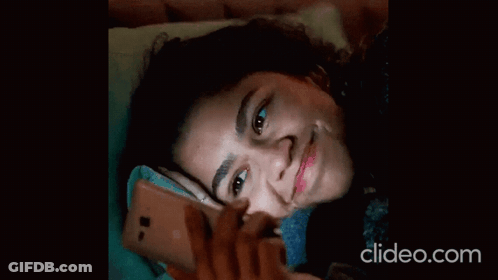 A Woman Laying On A Bed Looking At Her Phone With Clideo.com Written On The Bottom GIF