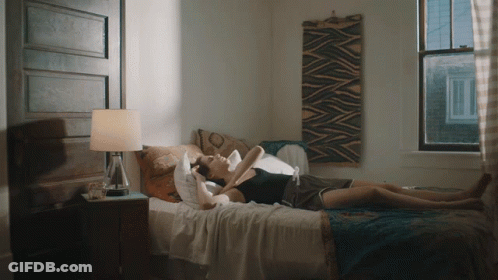A Woman Laying On A Bed With A Lamp And A Window Behind Her GIF