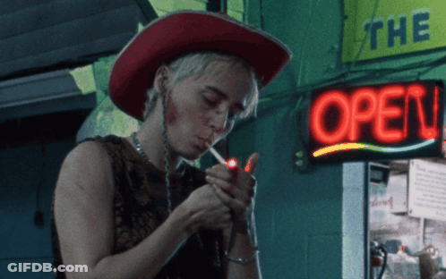 A Woman Lighting A Cigarette In Front Of A Neon Sign That Says Open GIF