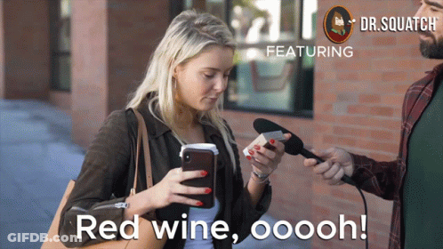 A Woman Looks At Her Phone While A Man Holds A Microphone And Says Red Wine Ooooh GIF