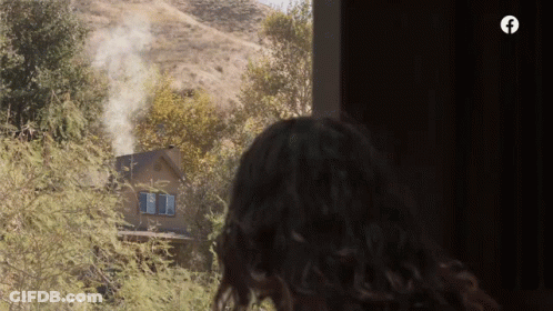 A Woman Looks Out A Window At A House With Smoke Coming Out Of It GIF