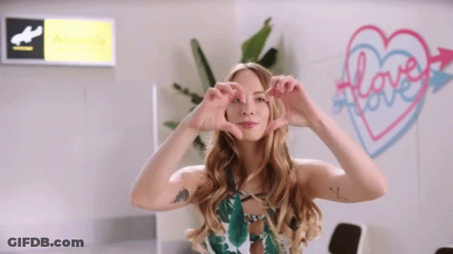 A Woman Making A Heart Shape With Her Hands In Front Of A Sign That Says Love GIF