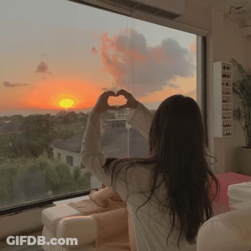A Woman Making A Heart Shape With Her Hands While Looking Out A Window At A Sunset GIF