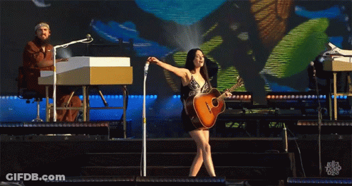 A Woman Playing A Guitar On Stage With A Man Playing A Keyboard In The Background GIF