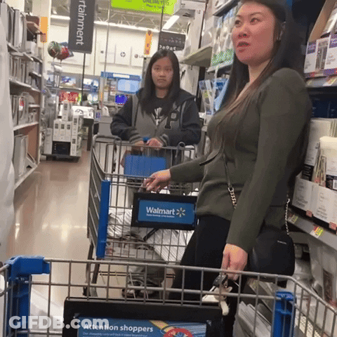 A Woman Pushing A Shopping Cart In A Store With A Sign That Says Attention Shoppers GIF