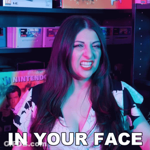 A Woman Says In Your Face While Standing In Front Of A Shelf Full Of Video Games GIF