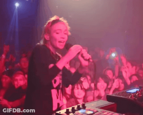 A Woman Singing Into A Microphone In Front Of A Crowd Wearing A Black Shirt That Says ' O ' On It GIF