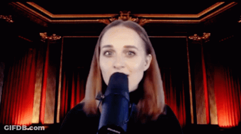 A Woman Singing Into A Microphone In Front Of A Red Curtain GIF