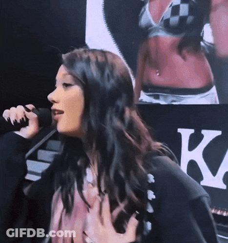 A Woman Singing Into A Microphone In Front Of A Sign That Says K. GIF