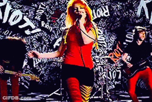 A Woman Singing Into A Microphone In Front Of A Wall With The Word Riot Written On It GIF