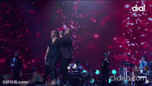 A Woman Singing Into A Microphone Next To A Man On A Stage With Dial Written On The Bottom GIF