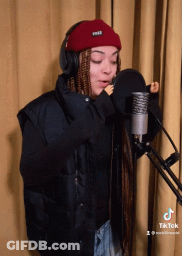 A Woman Singing Into A Microphone Wearing Headphones And A Red Beanie That Says Free GIF