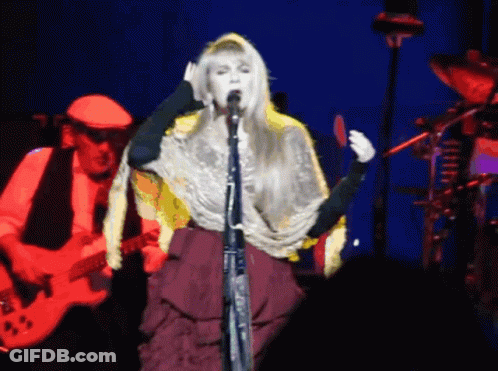 A Woman Singing Into A Microphone While A Man Plays A Guitar In The Background GIF