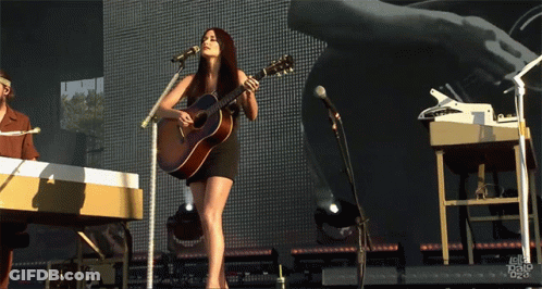 A Woman Singing Into A Microphone While Holding A Guitar On Stage GIF