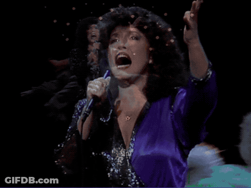 A Woman Singing Into A Microphone While Wearing A Purple Shirt GIF