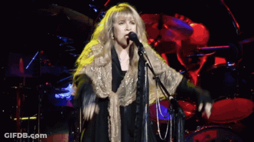 A Woman Singing Into A Microphone With A Drum Set In The Background GIF