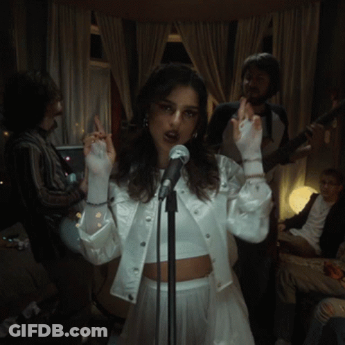 A Woman Singing Into A Microphone With A Man Playing A Guitar Behind Her GIF