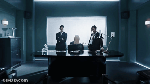 A Woman Sits At A Desk In A Dark Room With Two Men Standing Around Her GIF