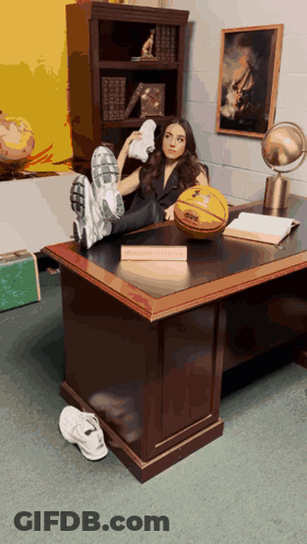 A Woman Sits At A Desk With Her Feet Up And A Basketball On The Desk With A Sign That Says Basketball Coach GIF