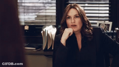 A Woman Sits At A Desk With Her Hand On Her Chin And A Picture Of A Police Officer Behind Her GIF