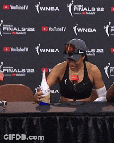 A Woman Sits At A Table In Front Of A Wall That Says Finals 22 On It GIF