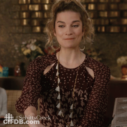 A Woman Sits At A Table With Her Hands On Her Chin While Holding A Cell Phone That Says #Schittscreek On It GIF