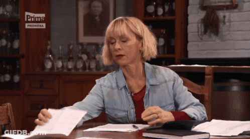 A Woman Sits At A Table With Papers In Front Of A Sign That Says Prima Premiera GIF