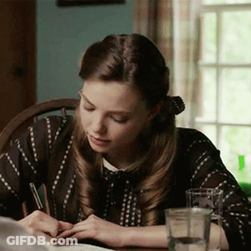 A Woman Sits At A Table Writing With A Pen GIF