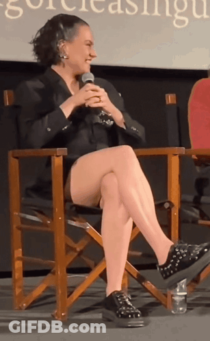 A Woman Sits In A Director 'S Chair With Her Legs Crossed And A Microphone In Her Hand GIF
