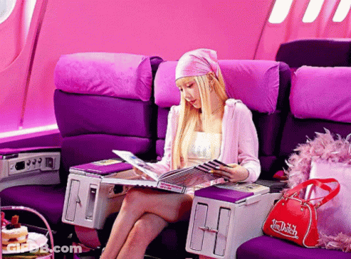 A Woman Sits In A Purple Chair Reading A Magazine While A Red Dutch Bag Sits Next To Her GIF